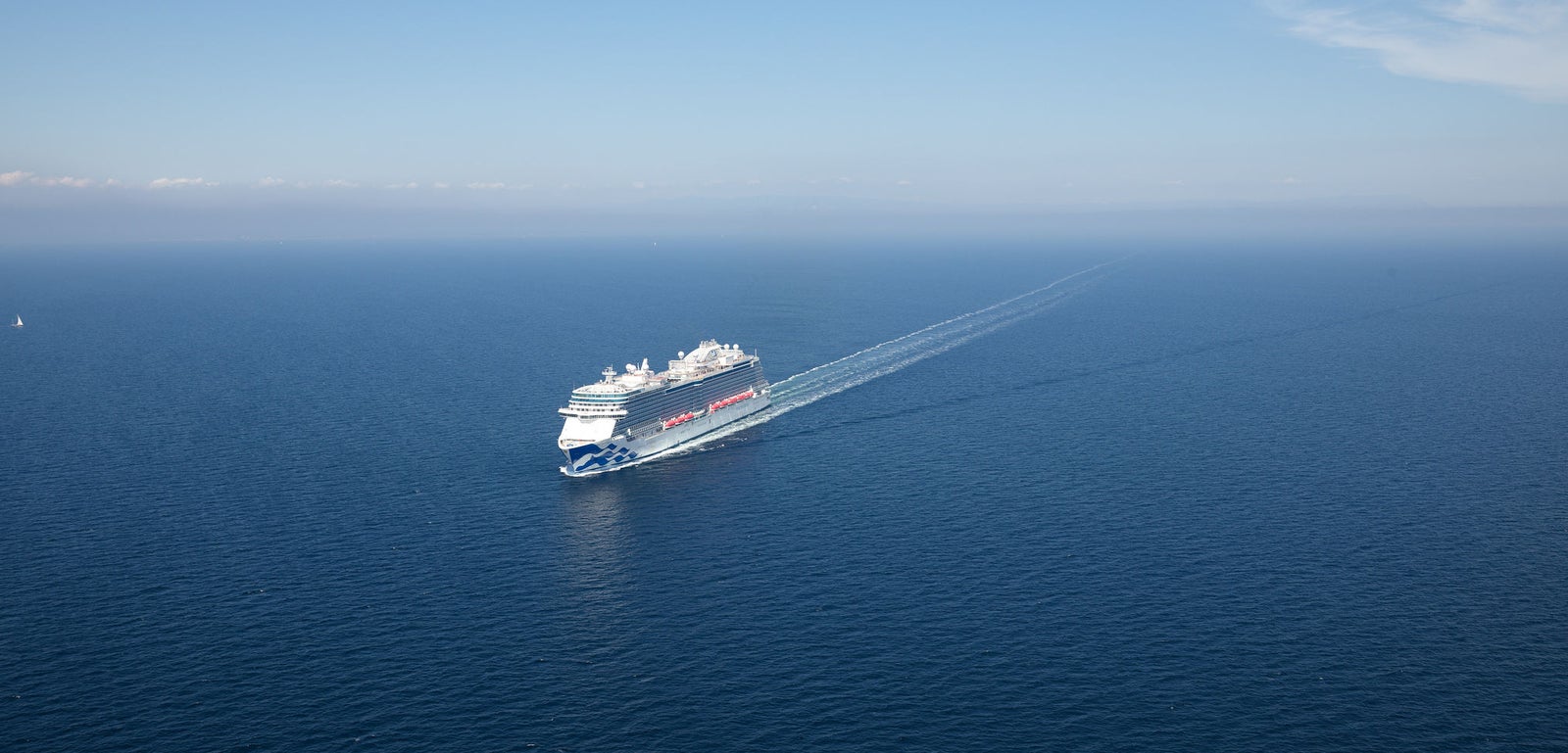 Fincantieri | Group - Business - Products - Cruise Ships - Enchanted Princess: Large operational cruise ship sailing on calm open waters under clear blue sky, white ship with visible red and blue accents, leaving wake in sea.