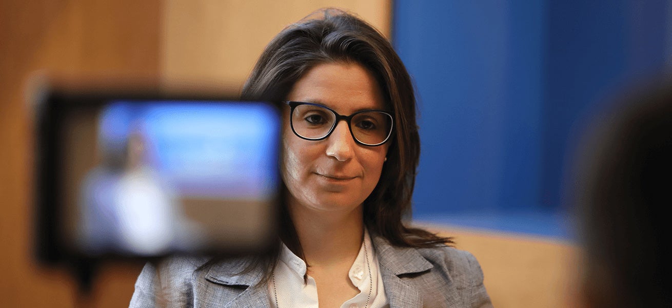 Fincantieri – People: Professional woman in a grey blazer and white shirt seated indoors, background with blue and wooden panels, camera in the foreground, formal and focused atmosphere, interview or recording setting.