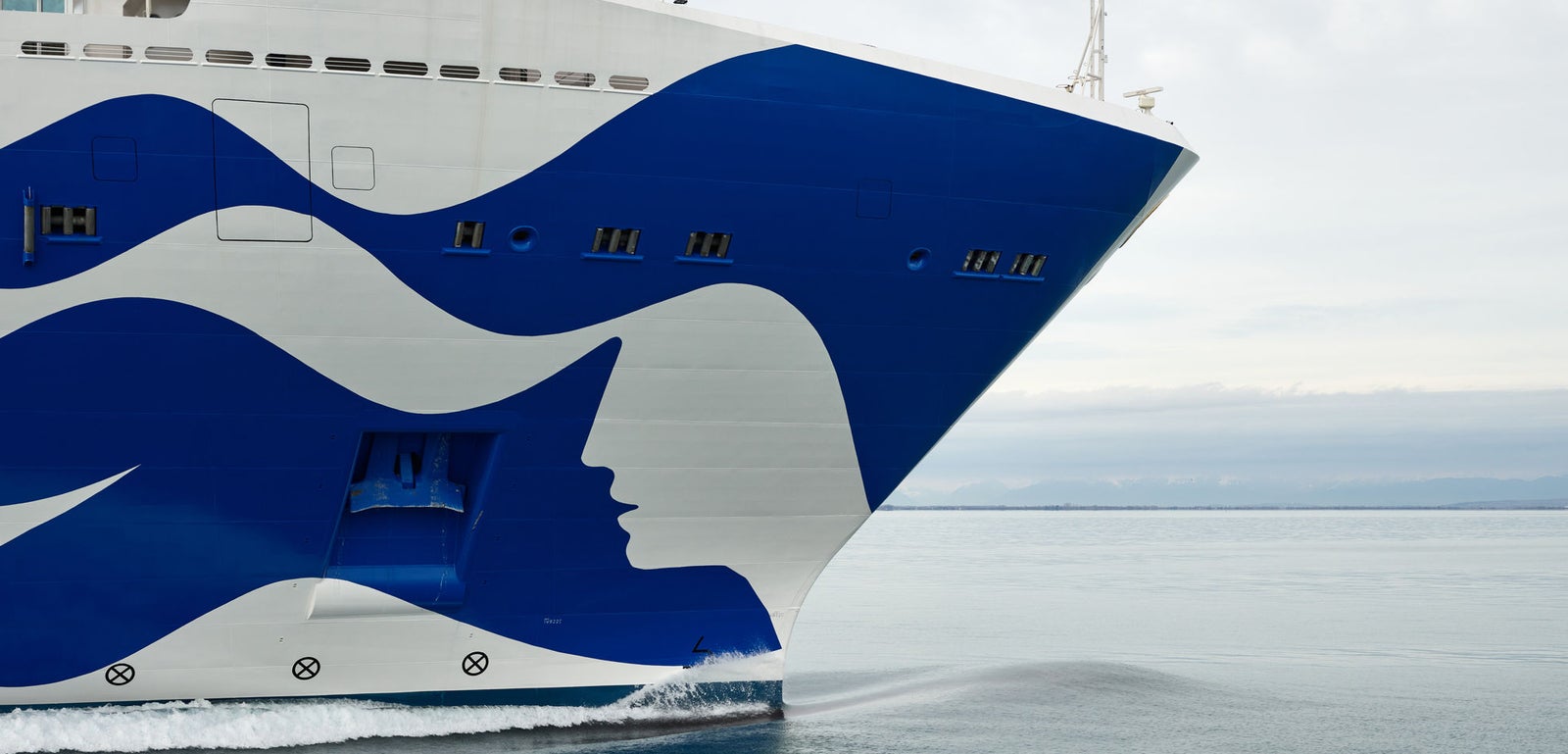 Fincantieri | Group - Business - Products - Cruise Ships - Discovery Princess: Close-up of Discovery Princess cruise ship bow in operation, blue wave design with face silhouette, gliding on calm waters under overcast sky.