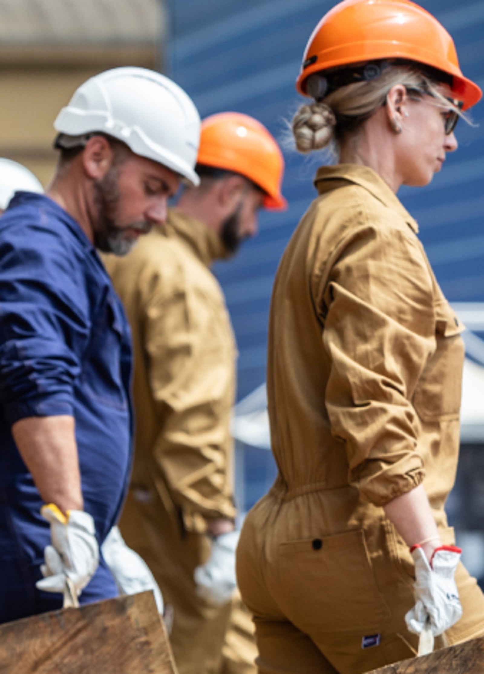 Fincantieri | People - Living Fincantieri - Diversity, Equity and Inclusion - DE&I Action Plan: A line of workers in tan jumpsuits and hard hats are walking outdoors. They appear to be part of a group or ceremony.