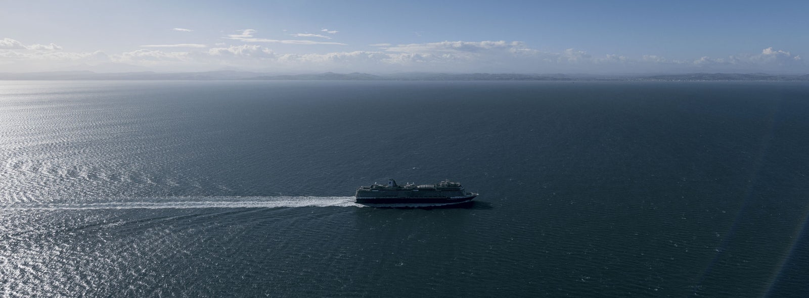 Fincantieri | Gruppo - Sostenibilità - Eccellenza industriale - Catena di fornitura: Grande traghetto operativo in navigazione su oceano calmo sotto cielo blu sereno, scia ben visibile e orizzonte lontano con profili di terra sullo sfondo.