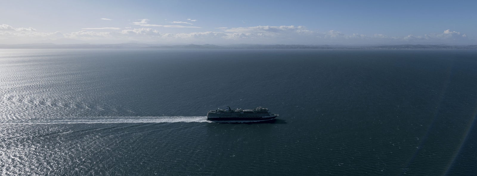 Fincantieri | Gruppo - Sostenibilità - Eccellenza industriale - Catena di fornitura: Grande traghetto operativo in navigazione su oceano calmo sotto cielo blu sereno, scia ben visibile e orizzonte lontano con profili di terra sullo sfondo.