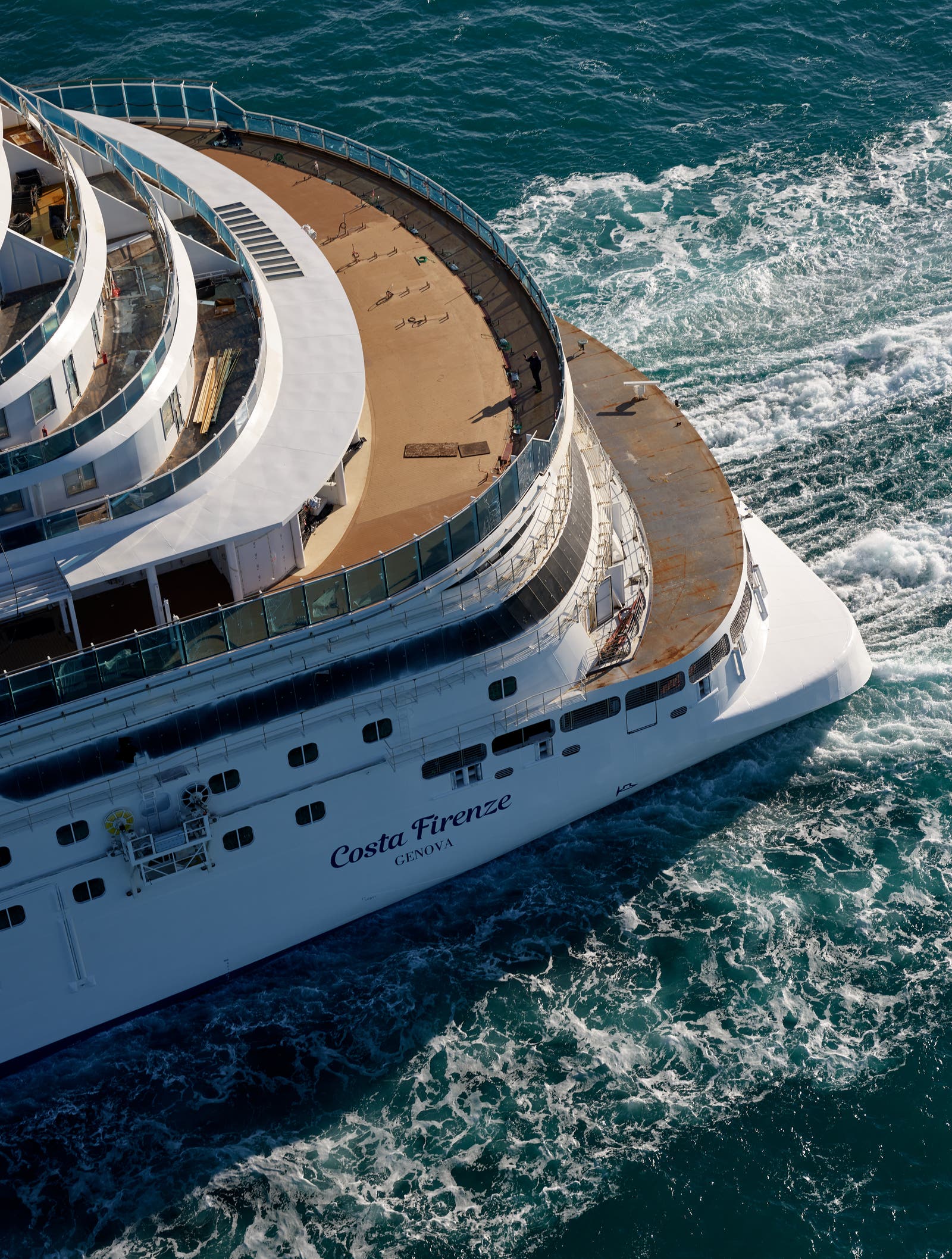 Fincantieri | Group - Business - Products - Cruise Ships - Costa Firenze: Rear view of Costa Firenze operational cruise ship, multiple decks visible, 'Costa Firenze' text on hull, sailing on vibrant blue waters with visible wake, serene maritime setting.