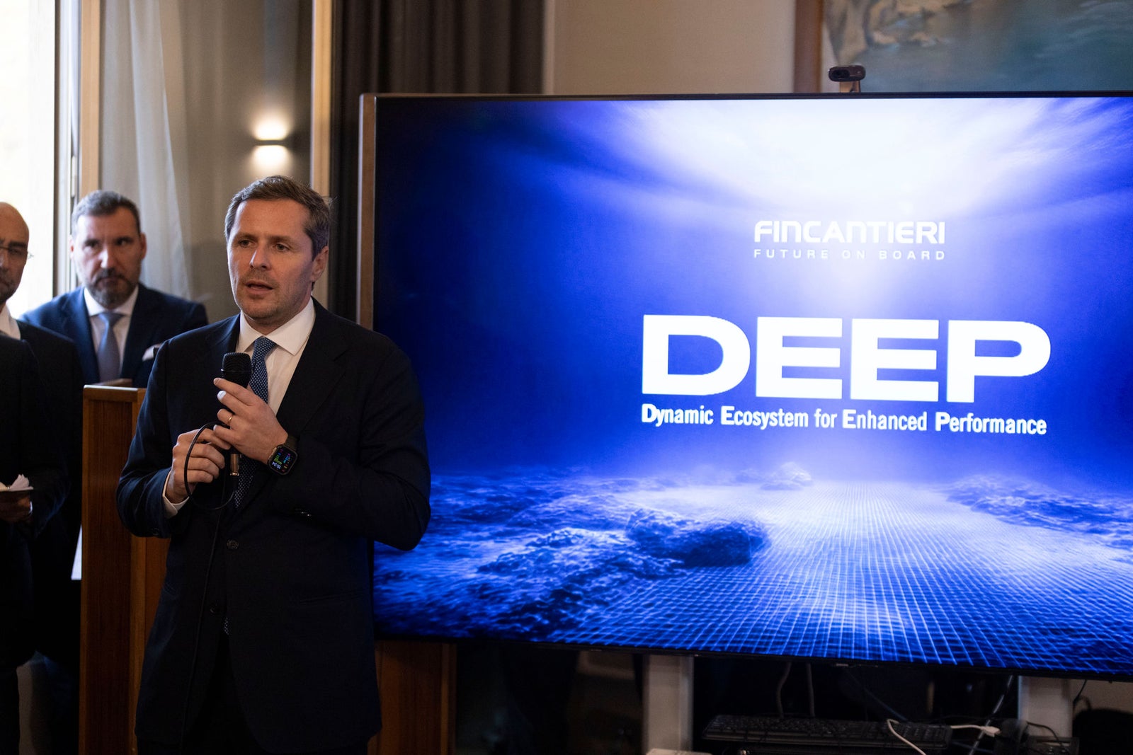 Fincantieri | Newsroom, Deep: A formal business presentation featuring a speaker in a suit holding a microphone. The setting appears to be an indoor corporate event with a large digital screen displaying the text 'DEEP' and 'Dynamic Ecosystem for Enhanced Performance'. The screen shows a futuristic blue and white design resembling a digital grid over Earth.