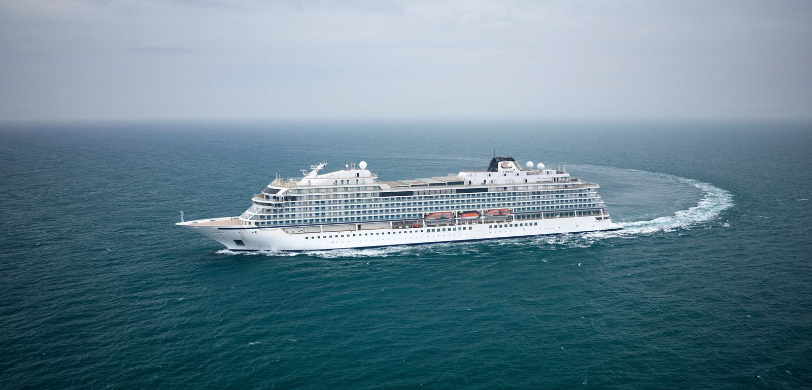 Fincantieri | Group - Business - Products - Cruise Ships - Viking Mars: Large white cruise ship in operation, navigating open ocean under cloudy sky, surrounded by calm waters, multiple decks and visible red lifeboats, emphasizing safety and luxury.