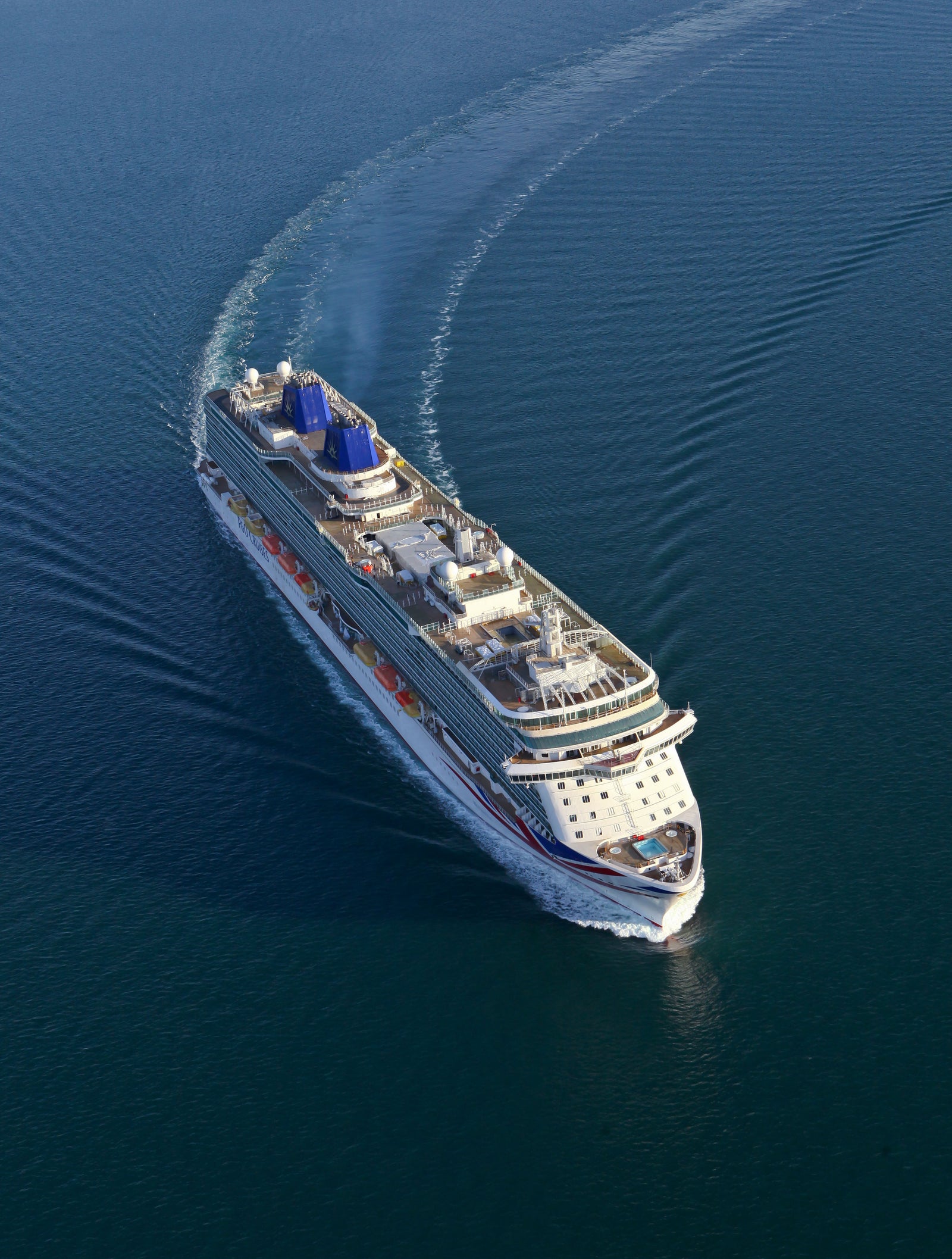 Fincantieri | Group - Business - Products - Cruise Ships - Britannia: Aerial view of Britannia cruise ship in operation navigating open sea, multiple passenger decks, visible outdoor areas, blue funnels, well-defined wake in water.