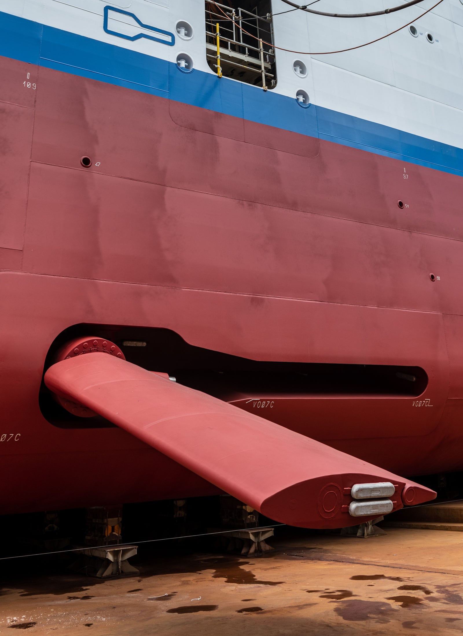 Fincantieri | Group - Business - Products - Stabilization, Propulsion, Positioning, and Generation Systems - Ship hull construction: Large red and white ship hull shown at an angle with red fin extended from side; ship in dry dock or under construction.