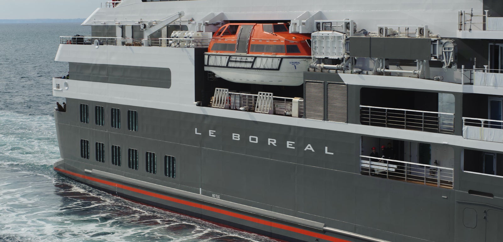 Fincantieri | Group - Business - Products - Cruise Ships - Le Boreal: Close-up of Le Boreal cruise ship in operation, dark grey hull with visible white text, orange and white lifeboat on upper deck, waves visible beneath hull.