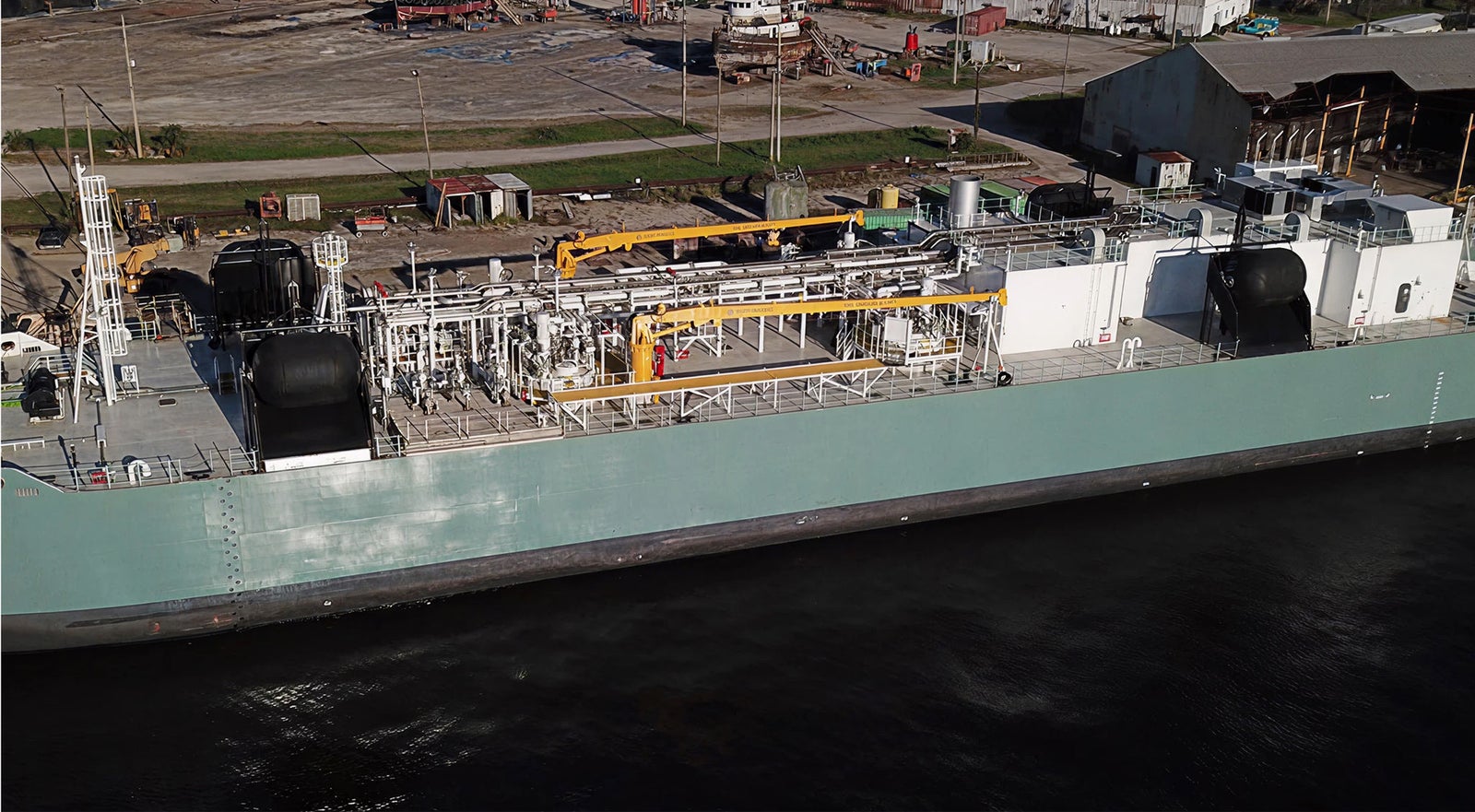 Fincantieri | Group - Companies - Subsidiaries and Affiliates - Fincantieri Marine Repair: Large industrial cargo ship docked in port, complex machinery and equipment on the deck, light green hull and visible piping systems; background with industrial buildings and storage areas along the quay.