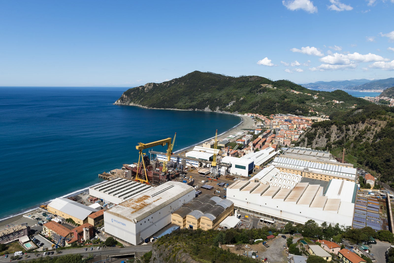 Fincantieri | Prodotti - Sistemi, componenti e infrastrutture - Complesso cantieristico: Vista panoramica su grande cantiere navale costiero con magazzini estesi, gru gialla di grande dimensione e navi ormeggiate; piccola città tra colline verdi e mare blu, cielo sereno. Nessun testo visibile.