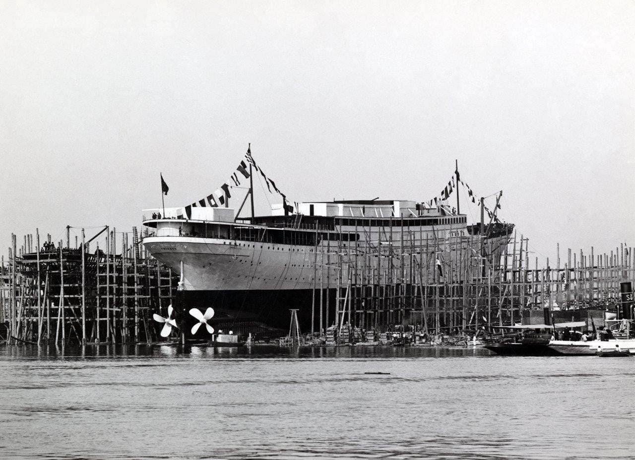 newsroom - press releases - 2026 - fincantieri group - fincantieri foundation: the first volume of the series on the history of italian shipbuilding now available in bookstores: fotografia storica in bianco e nero di una nave passeggeri in costruzione su pontili di legno e bandiere, vista dal porto.