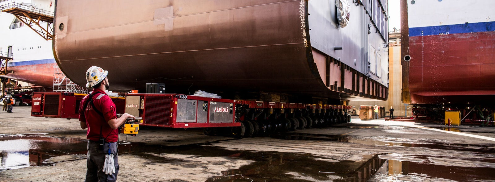 Fincantieri | Business Services - After-Sales Services - Lifecycle Management: A worker wearing protective gear supervises the transportation of a large ship hull in a shipyard. The setting features industrial equipment and wet ground, indicating recent activity or weather conditions. The hull is mounted on a heavy-duty transport vehicle, showcasing the scale of the operation. The worker holds a control device, emphasizing precision and safety.