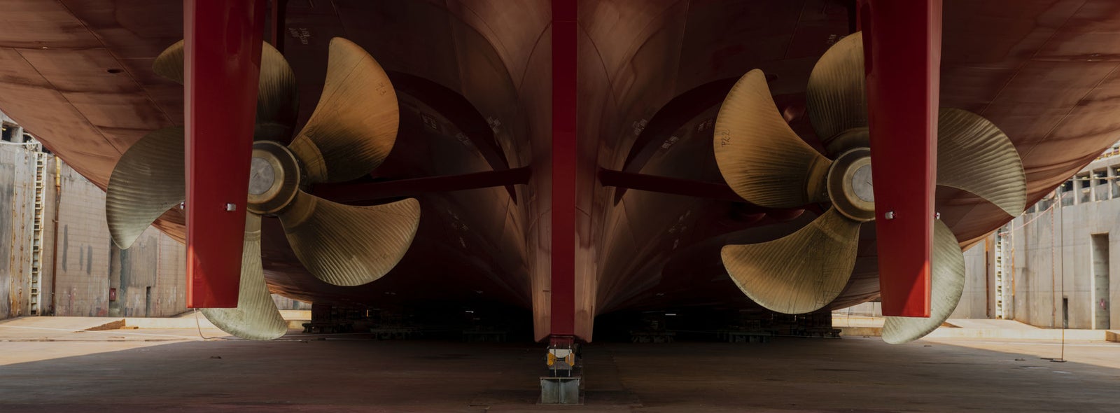 Fincantieri | Business Services - After-Sales Services - Lifecycle Management: Close-up view of two massive ship propellers beneath a vessel in a dry dock setting. The image highlights the metallic blades and the red hull of the ship, emphasizing industrial engineering and maritime design. The dry dock environment is visible, showcasing structural details and maintenance context.