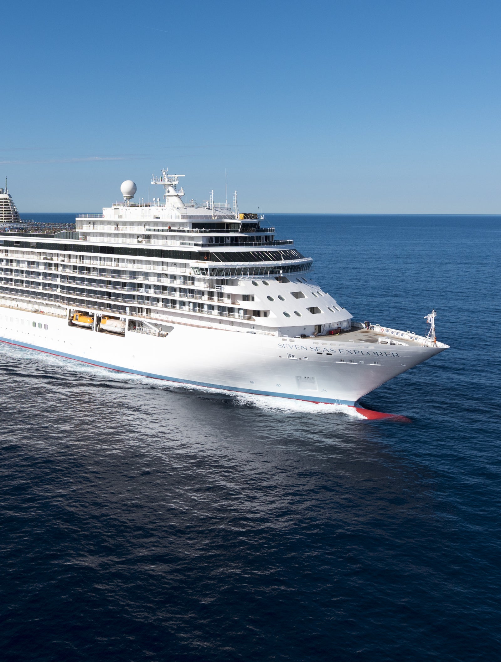 Fincantieri | Gruppo - Business - Prodotti - Navi da Crociera - Seven Seas Explorer: Grande nave da crociera moderna operativa bianca con molteplici ponti e scialuppe di salvataggio visibili, solca acque calme sotto cielo limpido, esperienza di viaggio di lusso con ampie viste sull’oceano e ambiente marittimo sereno e raffinato.