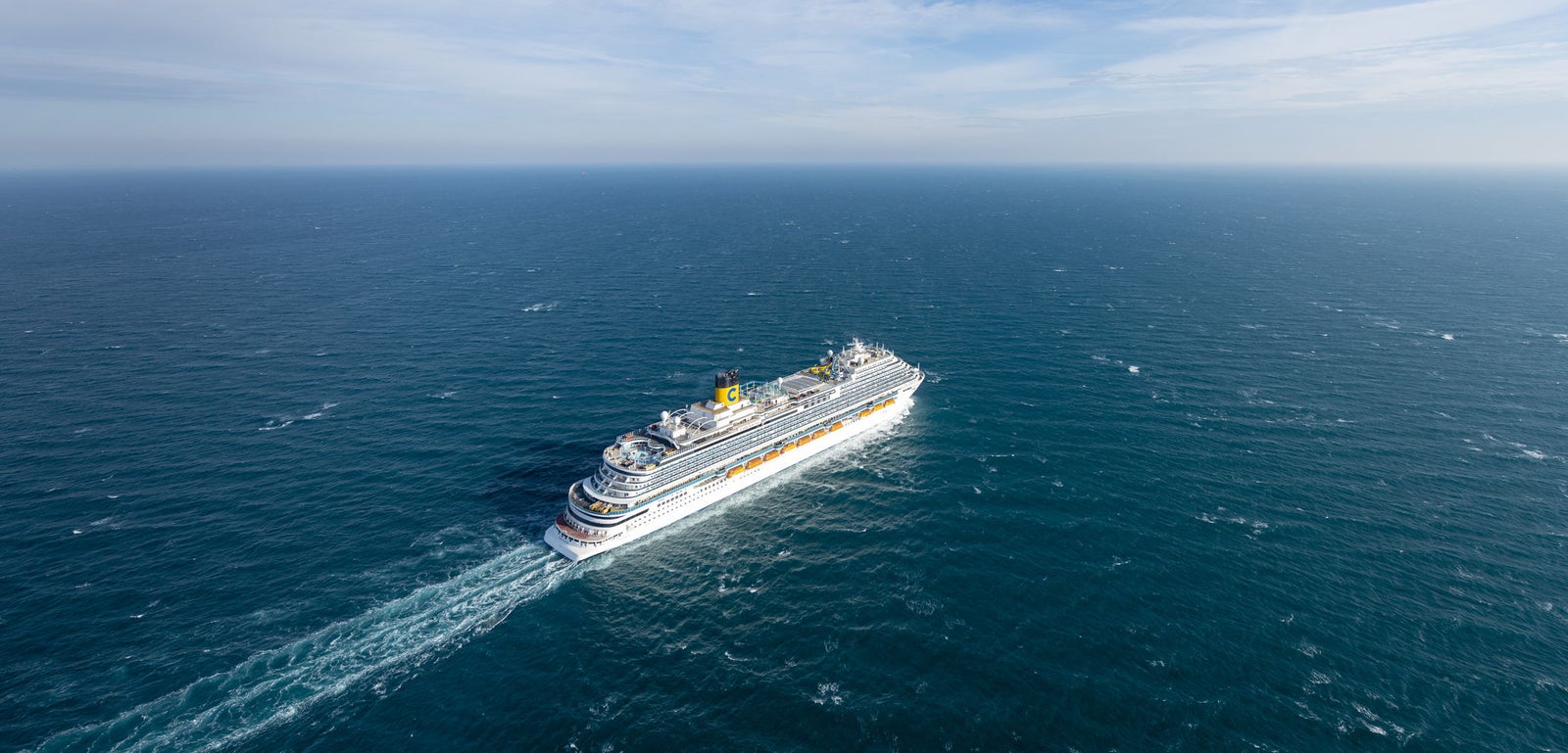 Fincantieri | Group - Business - Products - Cruise Ships - Costa Venezia: Large cruise ship in operation navigating vast open ocean under bright blue sky, surrounded by calm waters, white clouds streak across sky, multiple decks and visible structural details emphasize grandeur.