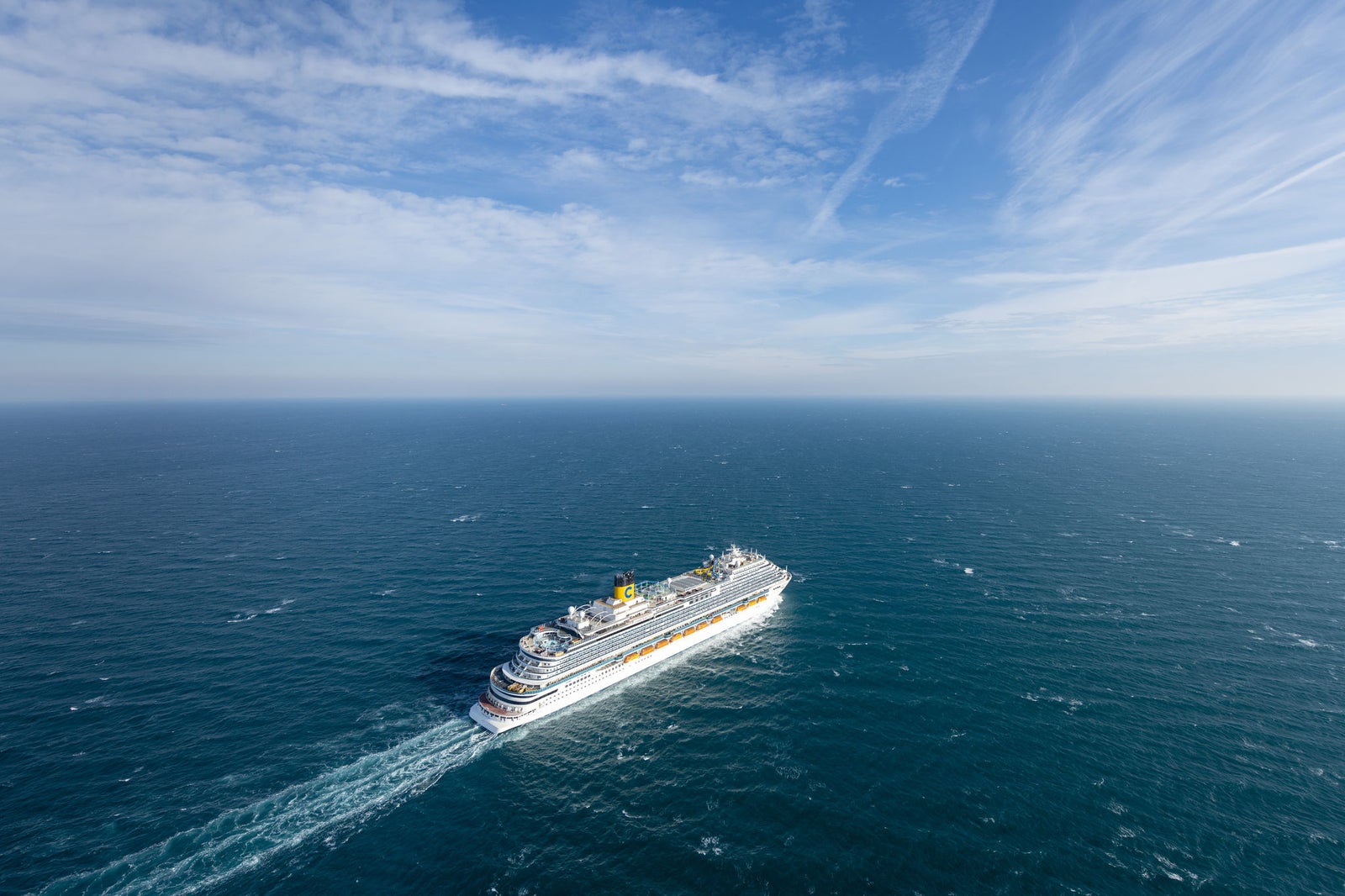 Fincantieri | Group - Business - Products - Cruise Ships - Costa Venezia: Large cruise ship in operation navigating vast open ocean under bright blue sky, surrounded by calm waters, white clouds streak across sky, multiple decks and visible structural details emphasize grandeur.