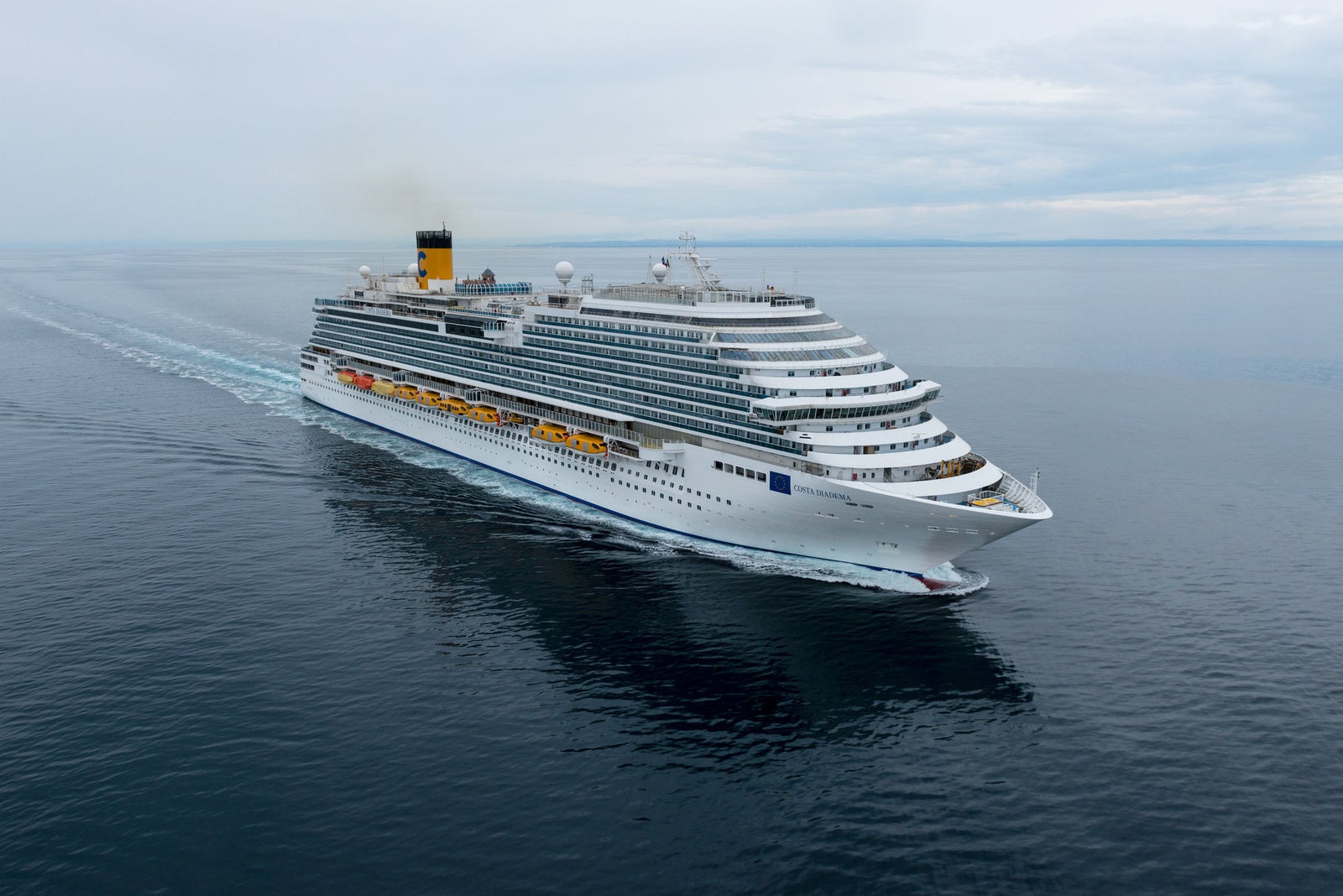 Fincantieri | Group - Business - Products - Cruise Ships - Costa Diadema: Massive cruise ship in operation gliding through calm open waters under cloudy sky, multiple decks, lifeboats, distinctive yellow funnel with visible markings, serene ocean setting highlights grandeur and scale, ideal for leisure travel and exploration.