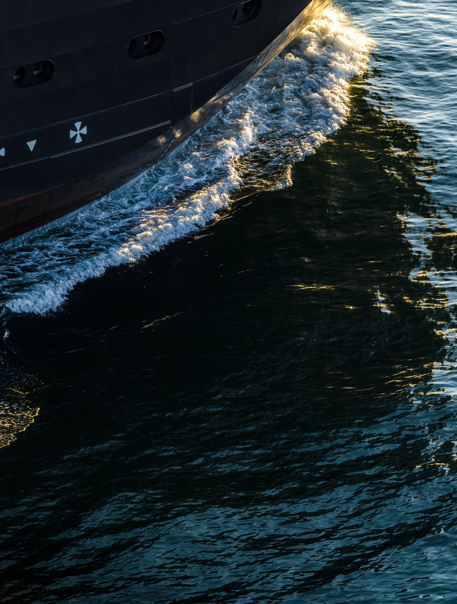 Prodotto - Nave da Ricerca Polare: Dettaglio prua della nave operativa in navigazione, scafo scuro, riflessi sull’acqua e increspature, atmosfera marittima tranquilla.