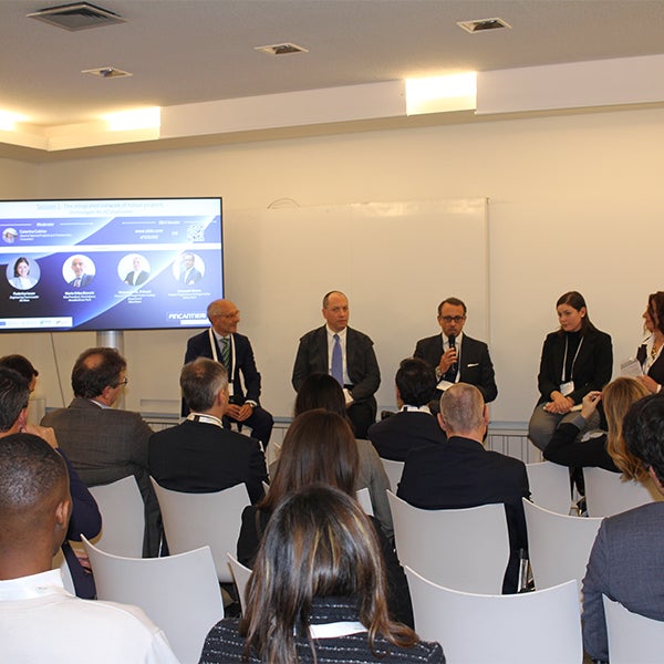 Fincantieri | Group - Innovation - Naval Ecosystem of the Future - Wave 2 the Future: Panel discussion with multiple speakers seated in a conference room, large screen displaying information and speaker visuals, audience arranged in rows, formal and business environment.