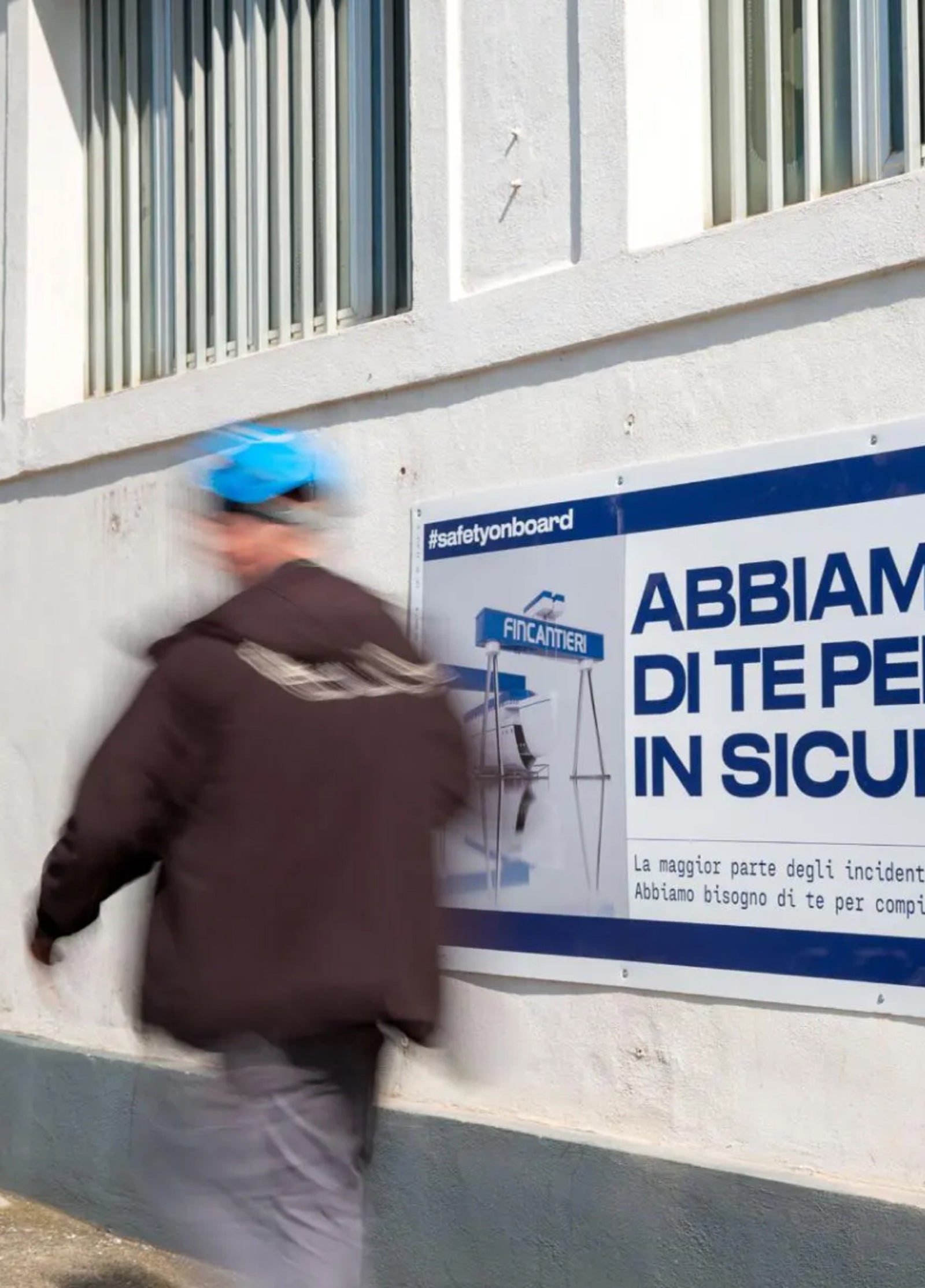 Fincantieri | People - Workplace Safety: Safety sign with the text “Abbiamo di te per in sicurezza” and hashtag #safetyonboard on a white wall, blurred figure of a worker wearing a blue cap and dark jacket in motion, industrial environment with large windows.