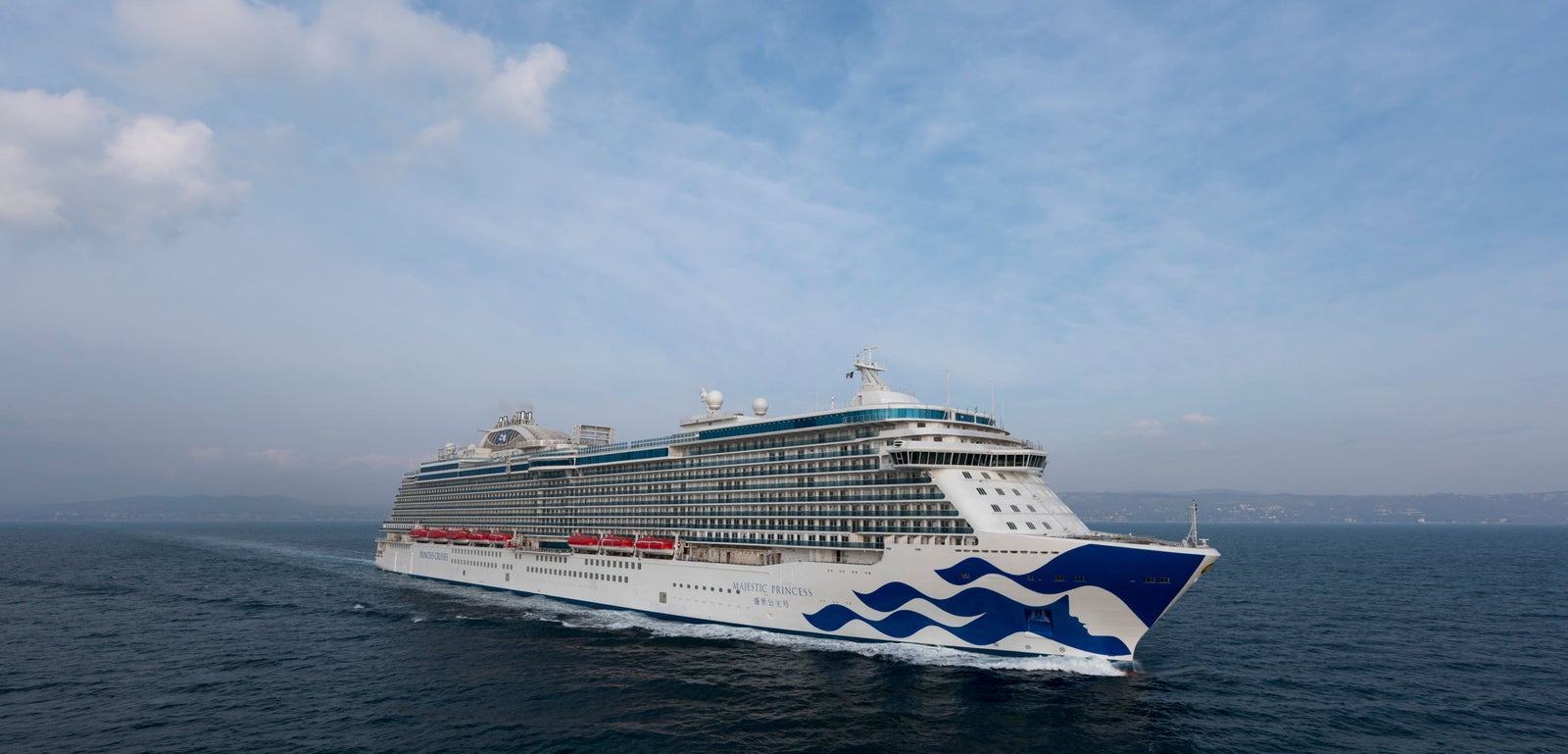 Fincantieri | Group - Business - Products - Cruise Ships - Majestic Princess: Side view of Majestic Princess Princess Cruises ship in operation, sailing on calm open waters under partly cloudy sky, multiple decks, red lifeboats and wave design on bow.