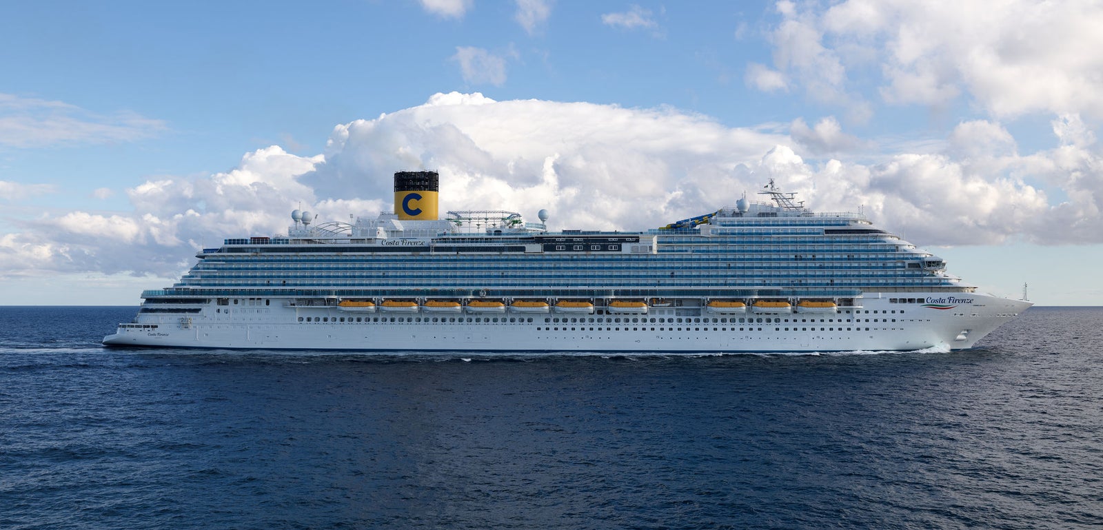 Fincantieri | Group - Business - Products - Cruise Ships - Costa Firenze: Large operational cruise ship with many decks and yellow funnel with 'C' logo, sailing on calm waters with land visible on horizon, imposing design reflecting grandeur and high passenger capacity.