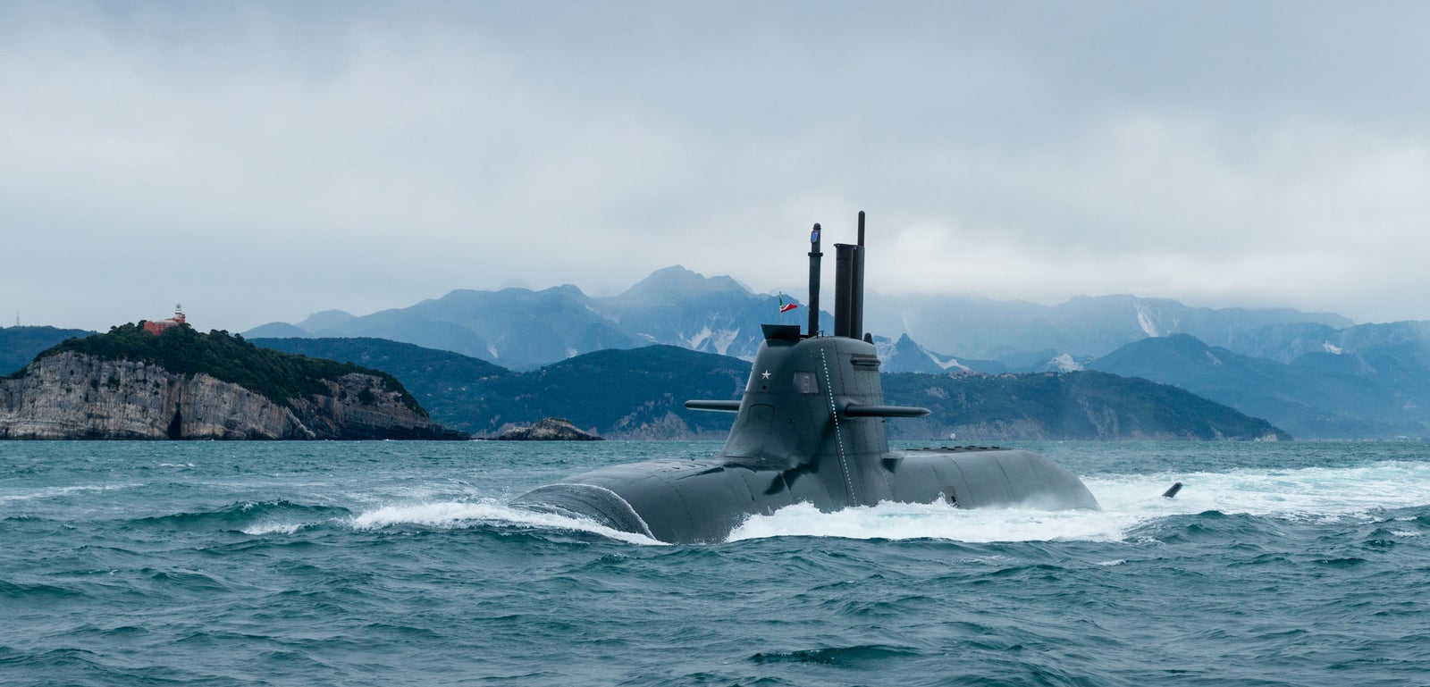 Fincantieri | Gruppo - Business - Prodotti - Sommergibili - Classe Todaro: Sommergibile moderno operativo, parzialmente immerso, design slanciato, in navigazione su acque oceaniche calme, costa montuosa sullo sfondo, cielo coperto.