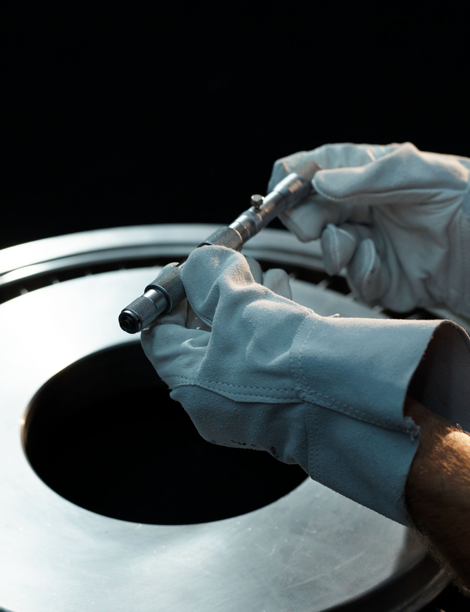 Fincantieri | Persone - Inclusione Vivere Fincantieri: Primo piano di mani con guanti che utilizzano uno strumento di misura su componente metallica di turbina, ambientazione industriale con luce focalizzata, attenzione alla precisione tecnica e alla professionalità.