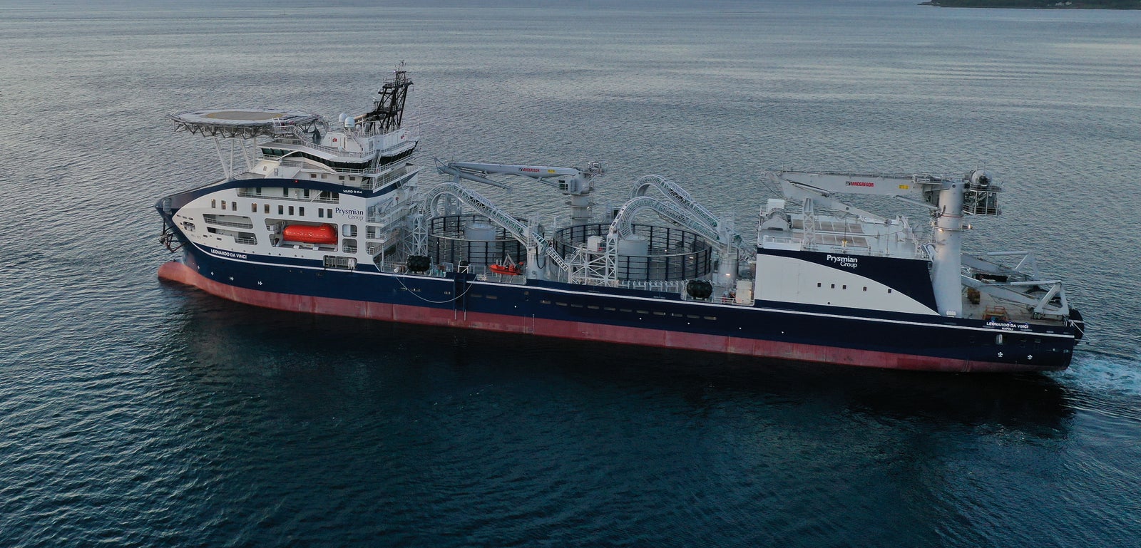 Prodotto - Nave Industriale Monna Lisa: Vista laterale della nave operativa in navigazione al tramonto, equipaggiamenti e strutture per operazioni marittime visibili, sfondo con montagne lontane e cielo tenue.