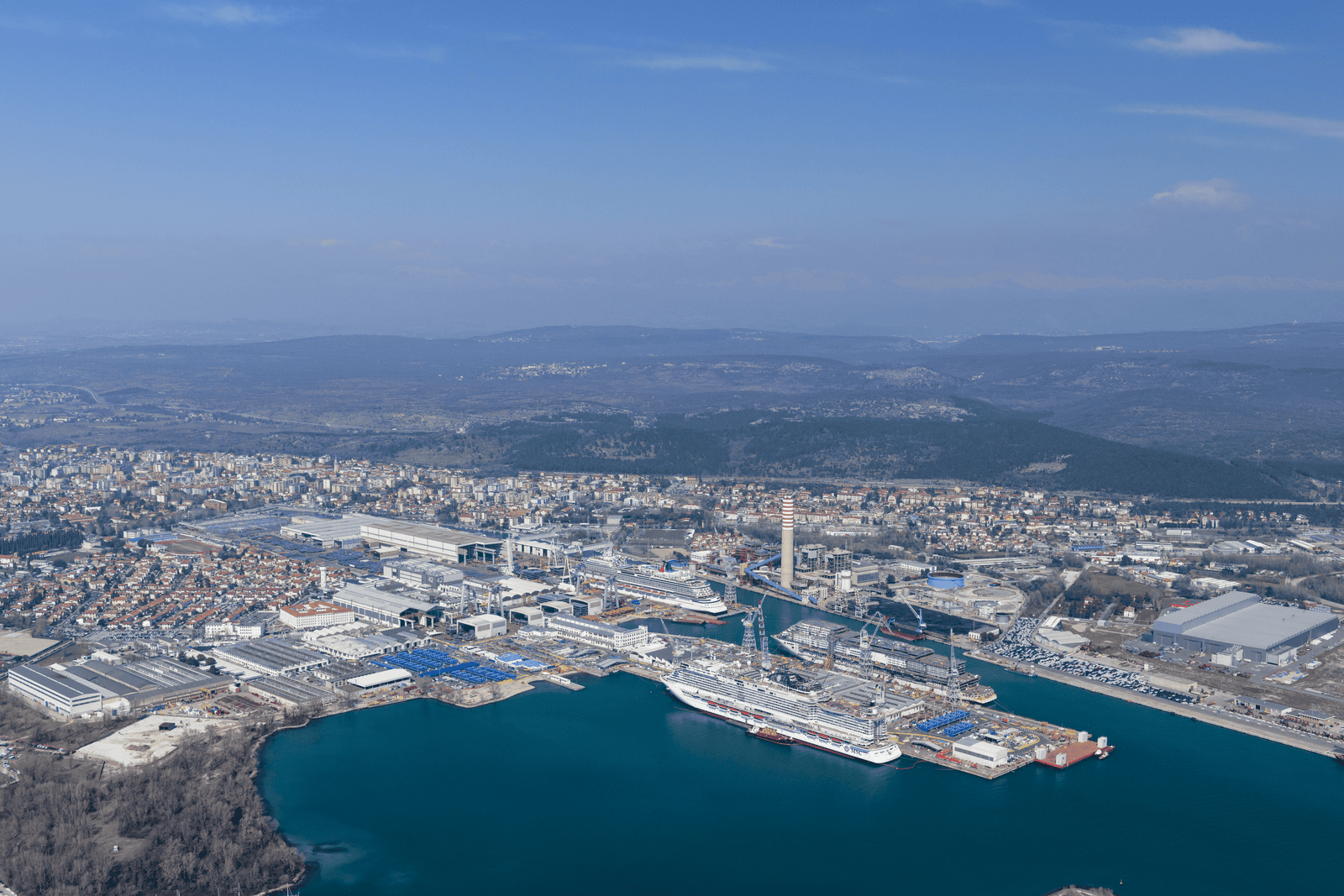 Fincantieri | People - Diversity, Equity and Inclusion: Aerial view of a busy industrial port near a coastal city, docked ships, warehouses and chimney, urban context and natural landscape, blue sky and calm waters.
