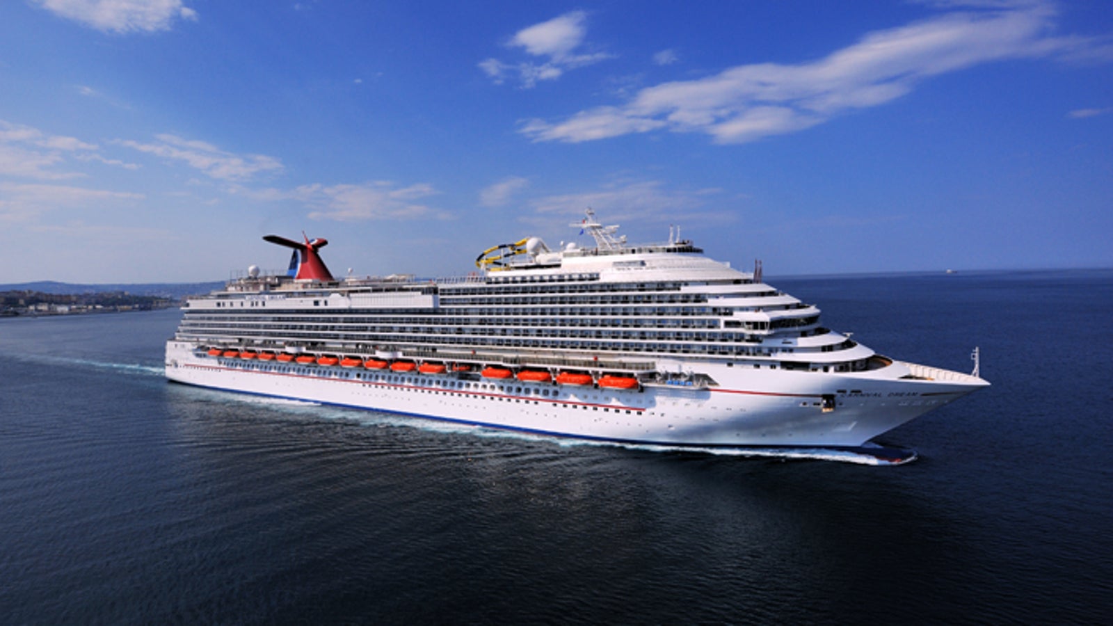 Fincantieri | Group - Business - Products - Cruise Ships - Carnival Dream: Large operational white cruise ship with multiple decks sailing on calm open waters under clear blue sky, red lifeboats and red wing-shaped funnel.