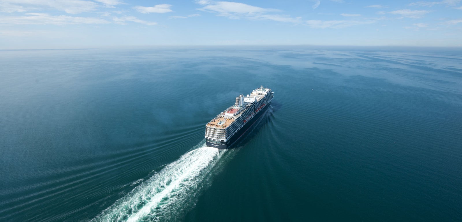 Fincantieri | Group - Business - Products - Cruise Ships - Nieuw Amsterdam: Large operational cruise ship sailing on calm open waters under clear blue sky, visible wake, smaller ship and cargo vessel distant on horizon.