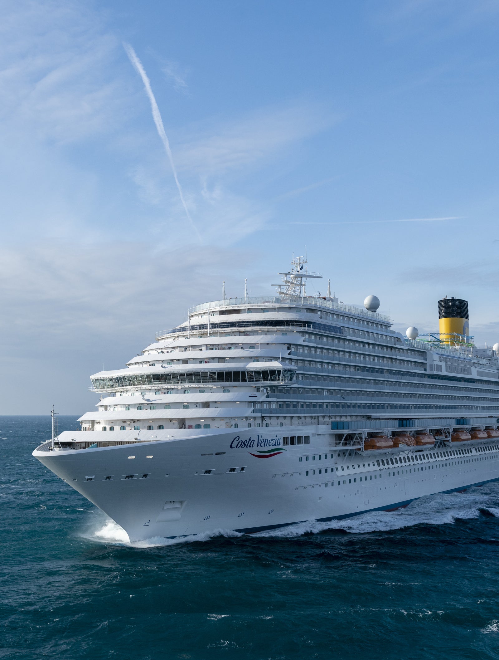 Fincantieri | Group - Business - Products - Cruise Ships - Costa Venezia: Large operational white cruise ship with multiple decks, visible lifeboats and distinctive yellow funnel, sailing on calm waters under clear sky, atmosphere of adventure and leisure.