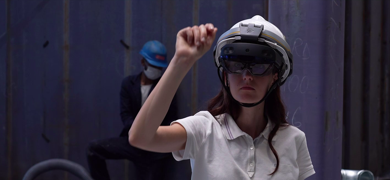 Fincantieri – People: Person wearing a white helmet and black polo shirt walking on a metal platform in an industrial environment, with steel structures, machinery, and blue sky in the background; active shipyard or production setting.