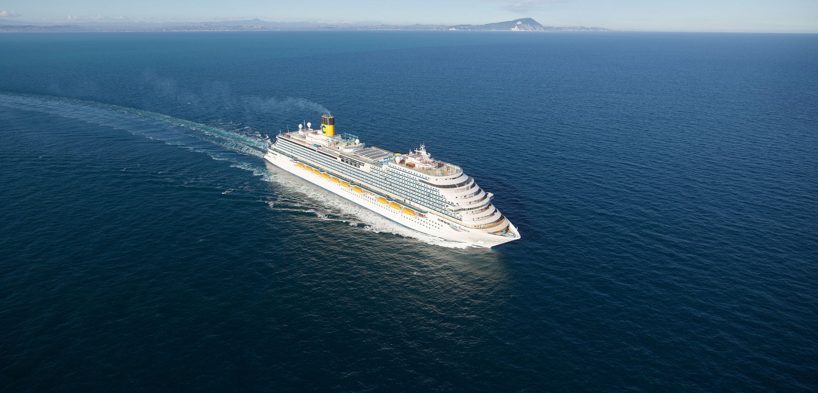 Fincantieri | Group - Business - Products - Cruise Ships - Costa Firenze: Large operational cruise ship with multiple decks and yellow funnel with 'C' logo, sailing on calm waters with land visible on horizon, imposing design highlighting the ship’s grandeur.