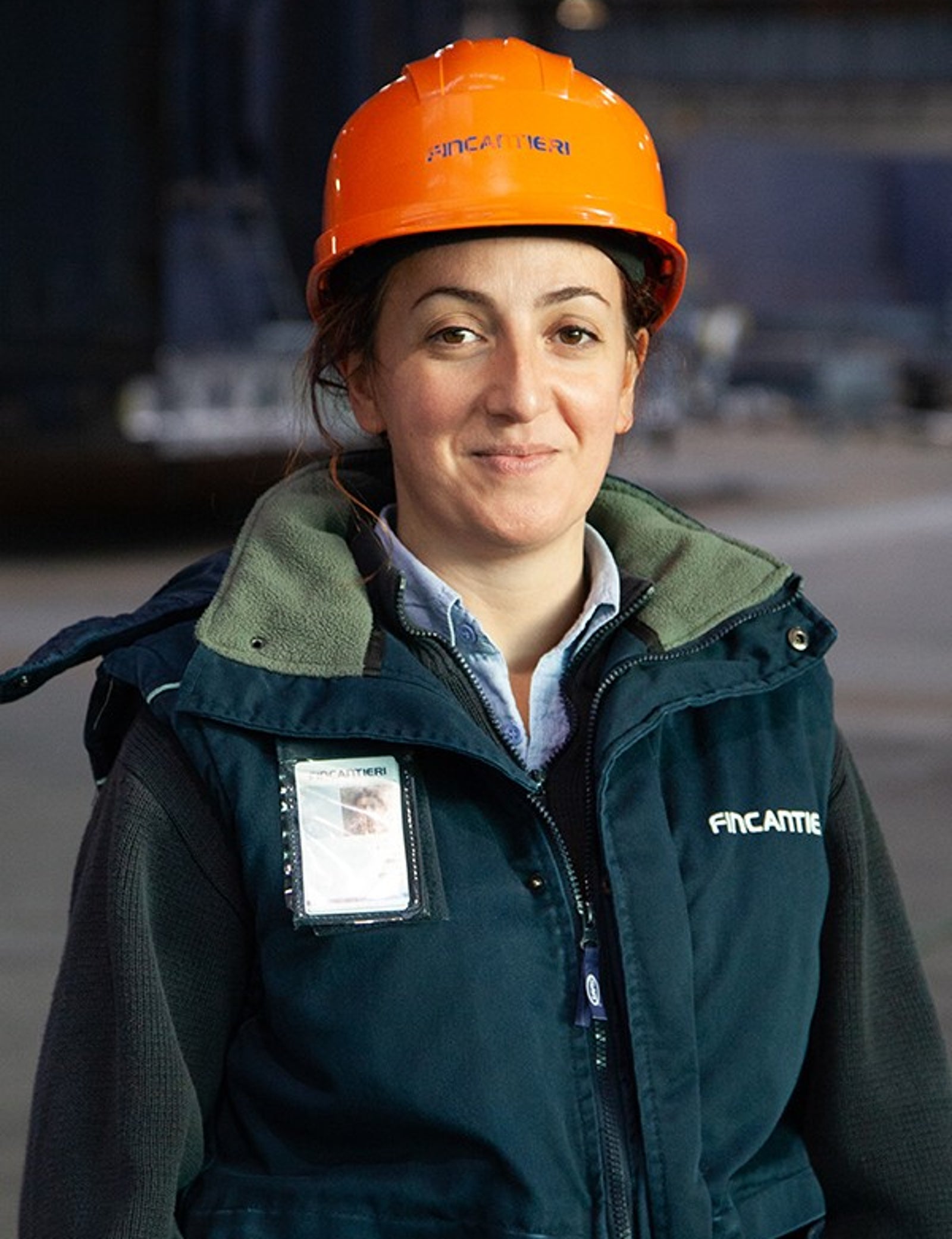 Fincantieri | Persone – Inclusione, Vivere in Fincantieri: Persona con casco arancione e gilet verde scuro in ambiente industriale, “FINCANTIERI” ben visibile sul casco, badge identificativo, sfondo con attrezzature industriali e illuminazione soffusa.