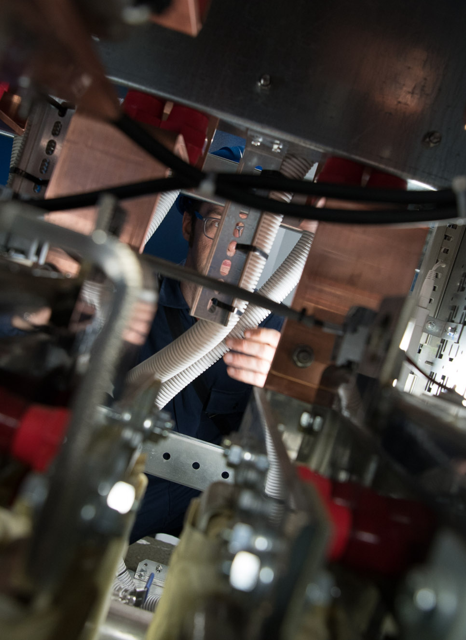 Fincantieri | Group - Business - Products - Systems Integration - Technician maintenance: Male technician with safety glasses and blue cap working on complex industrial machinery; manufacturing environment with visible cables and metallic components, hands-on maintenance or assembly in a technical workspace.
