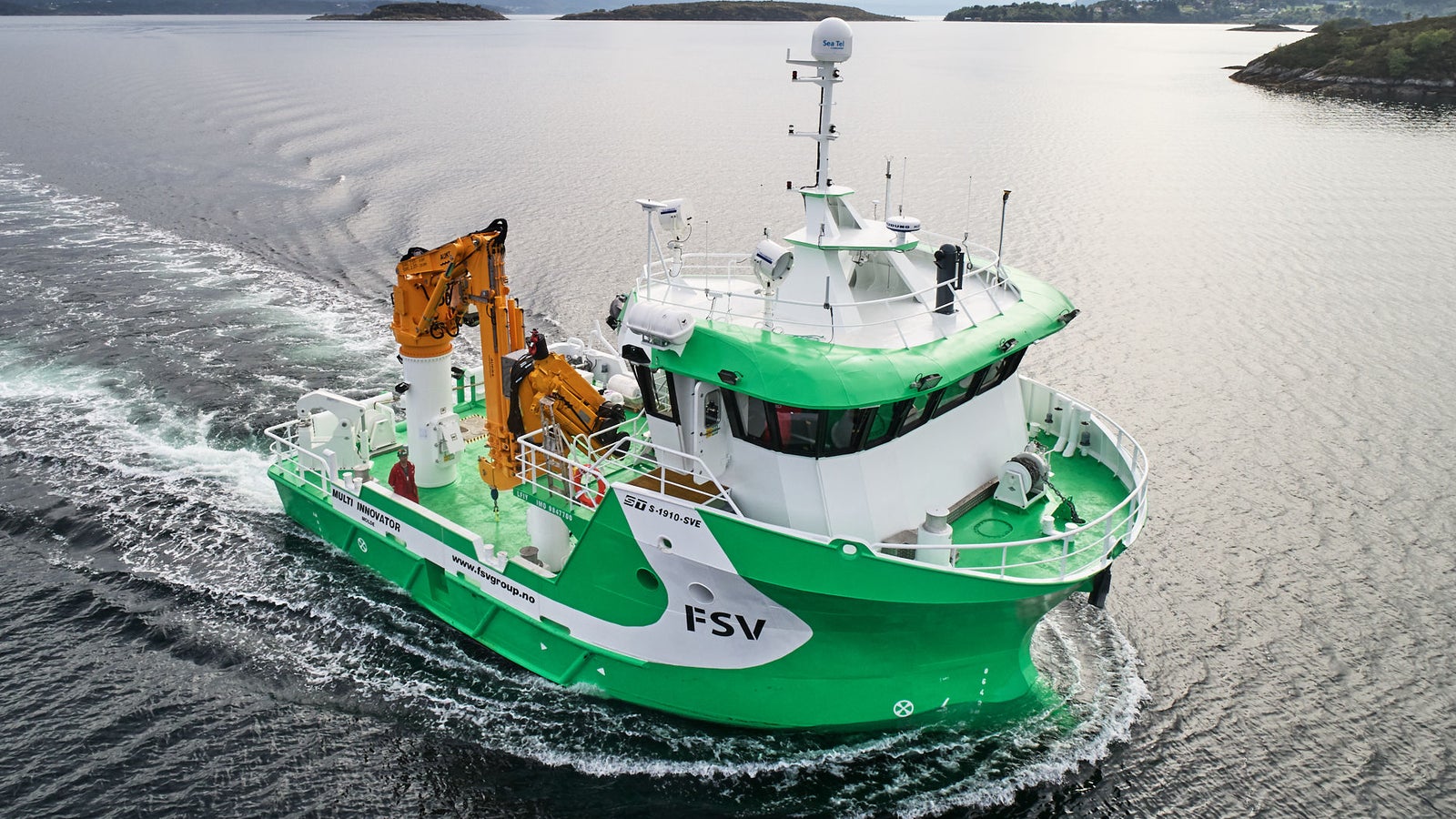 Prodotto - Nave Multi Innovator: Vista laterale della nave operativa in navigazione, scafo verde e bianco con scritta FSV, grande gru gialla visibile sul ponte, sfondo montuoso e cielo nuvoloso.