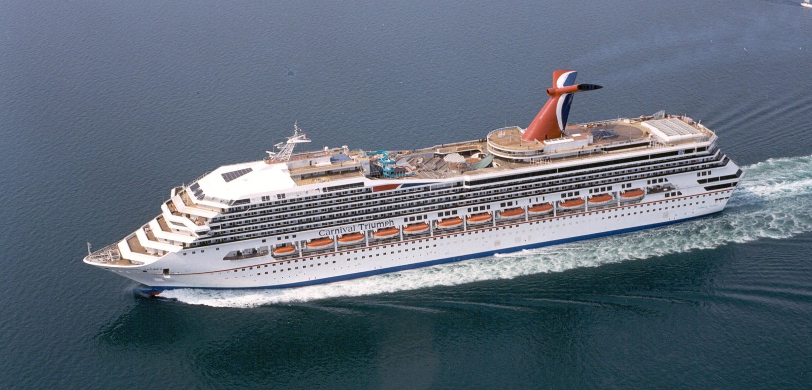 Fincantieri | Group - Business - Products - Cruise Ships - Carnival Triumph: Large operational cruise ship sailing on calm open waters, multiple decks including upper recreational areas, distinctive red and blue funnel clearly visible, white exterior and rows of windows.
