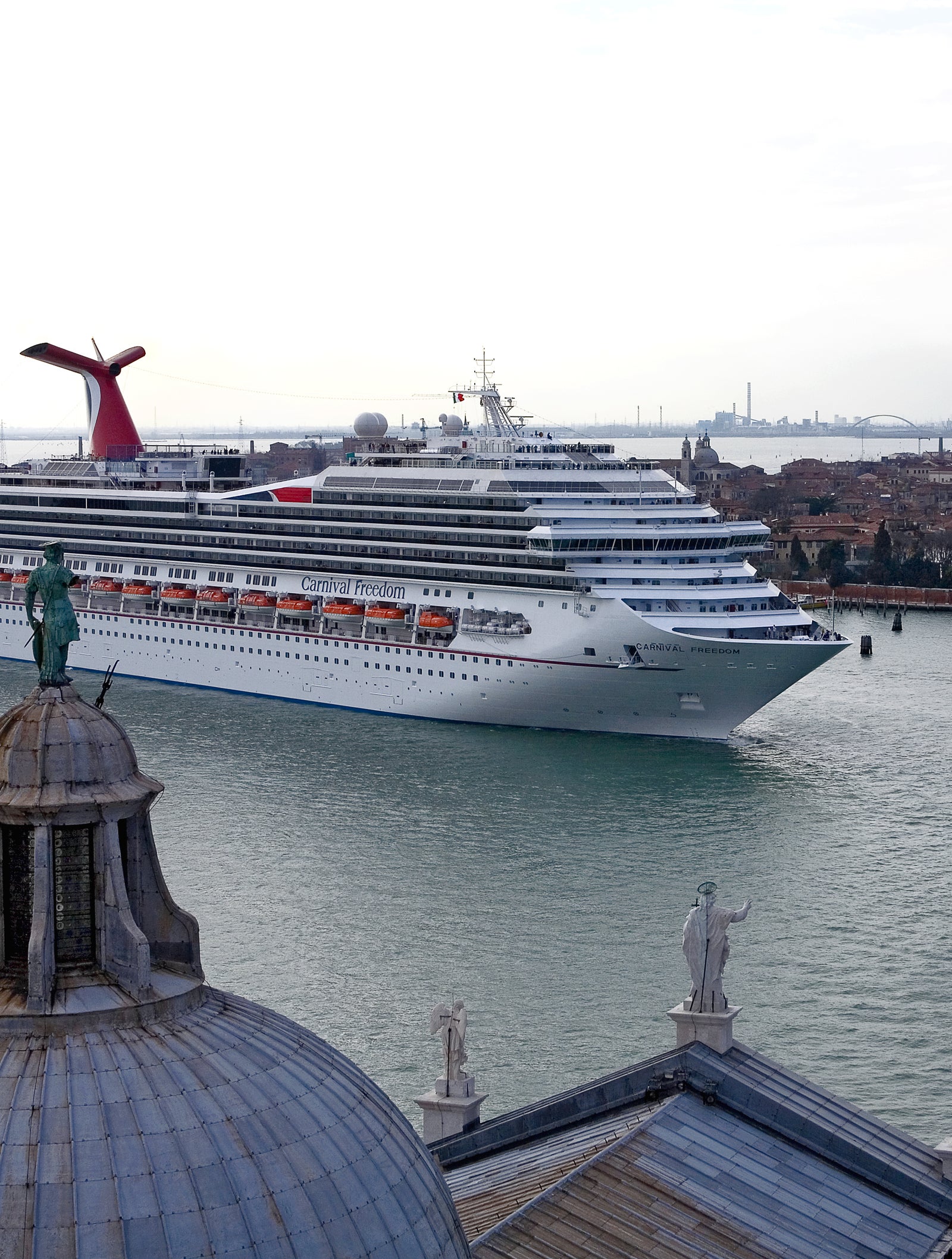 Fincantieri | Gruppo - Business - Prodotti - Navi da Crociera - Carnival Freedom: Grande nave da crociera operativa che naviga attraverso un canale veneziano, circondata da architettura storica e monumenti, in primo piano edificio a cupola con statue, sullo sfondo strutture iconiche veneziane e canali.