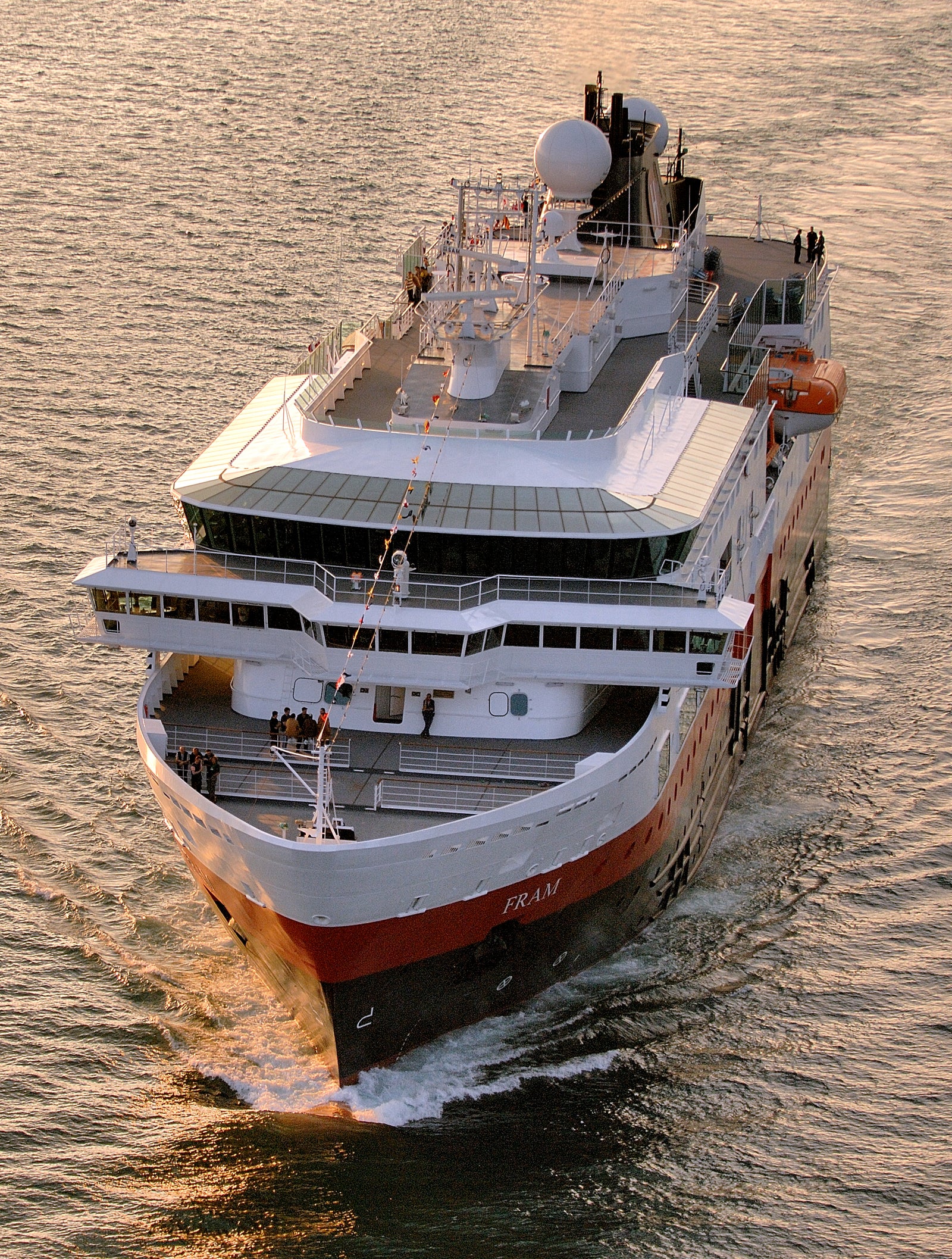 Fincantieri | Group - Business - Products - Cruise Ships - Fram: Imposing operational ship with red and white exterior, lifeboats and prominent radar dome on top, 'Fram' name clearly visible on hull, sailing on calm waters under golden sunset, serene and evocative maritime travel atmosphere.