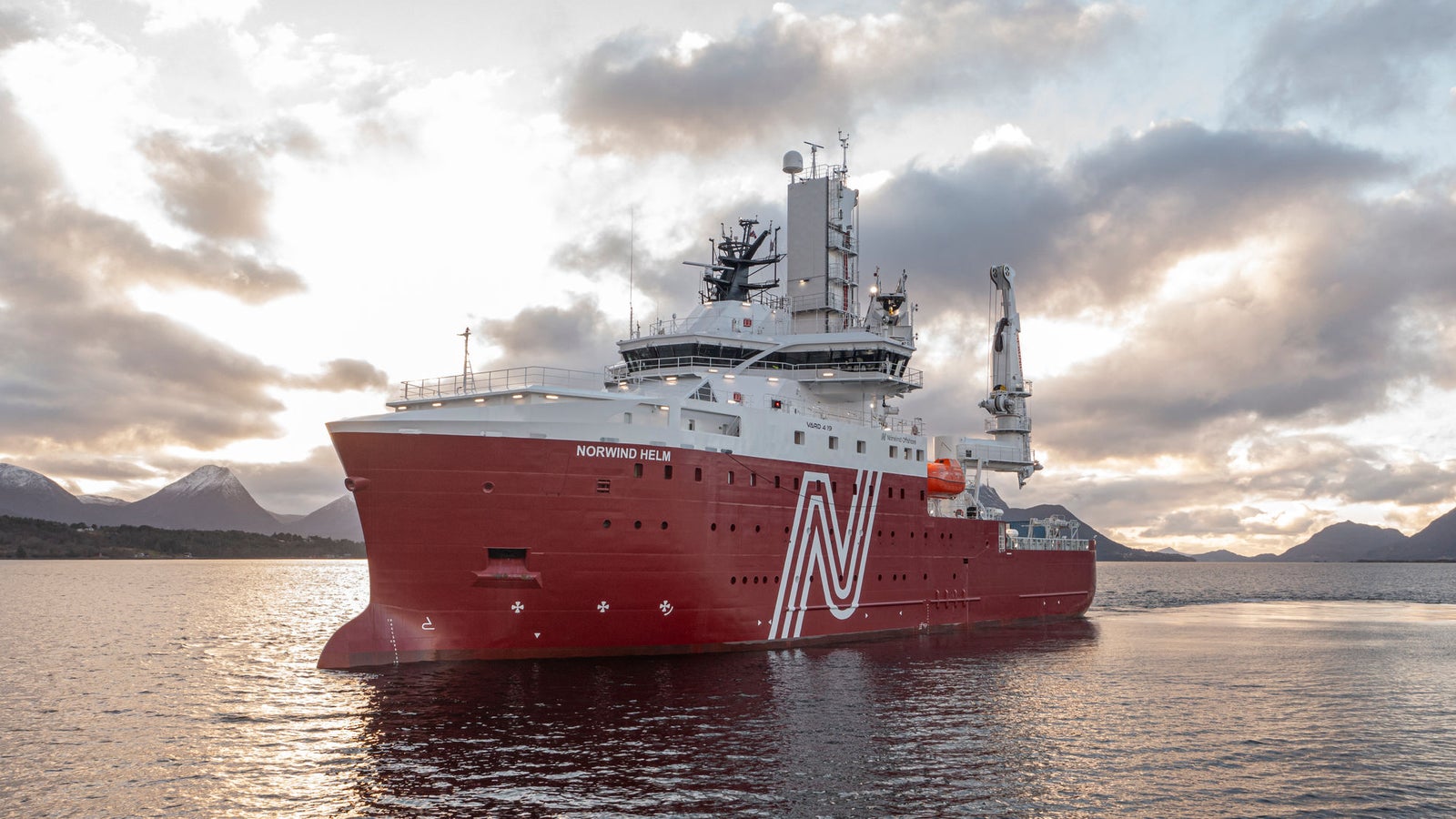 Confermato, grazie della correzione.  Prodotto - Offshore Support Vessel Norwind Helm: Vista laterale della nave operativa al tramonto, scafo rosso con logo N bianco e nome Northwind Helm visibile, gru e attrezzature marittime avanzate, sfondo con profilo montuoso e cielo parzialmente nuvoloso.