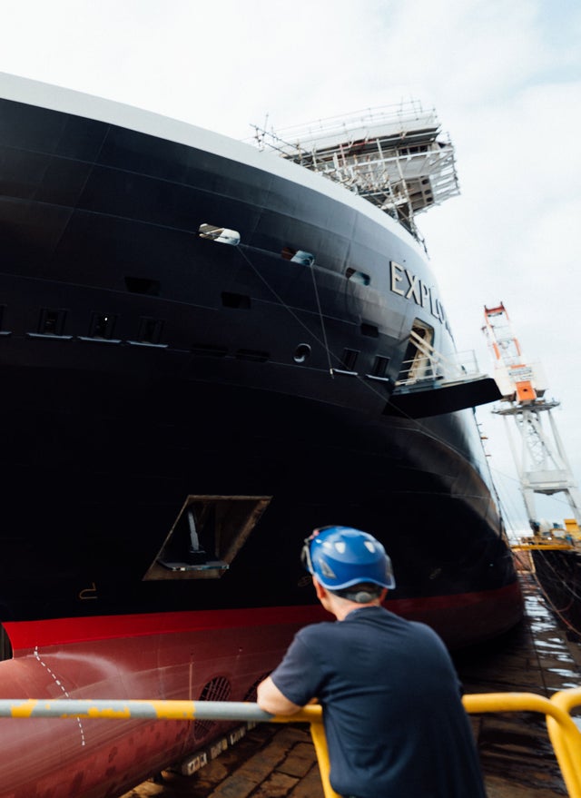 Fincantieri | Gruppo - Sostenibilità - Eccellenza industriale - Cilenti: Nave di grandi dimensioni con testo 'EXPL' visibile è ormeggiata in cantiere navale durante la costruzione; operaio con casco blu osserva dietro parapetto giallo, scafo nero e rosso con ponteggi sulla struttura superiore, ambiente industriale con gru e attrezzature sullo sfondo.