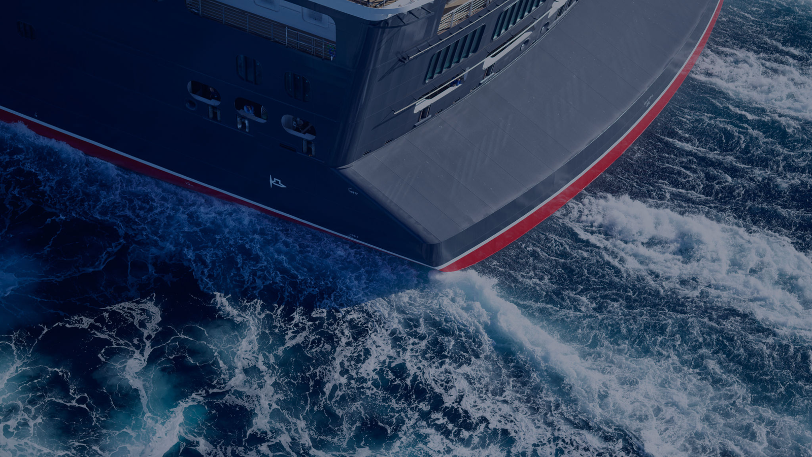 Fincantieri | Group - Sustainability - Energy Transition - Net-zero: Bow of a large ship with elegant design and red stripe on the hull, sailing through lively waves generating patterns of white foam, deep blue marine environment.