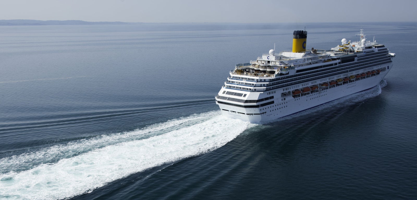 Fincantieri | Group - Business - Products - Cruise Ships - Costa Favolosa: Large white cruise ship with multiple decks in operation sailing through calm open waters, visible wake behind, yellow and black funnel, faint horizon in distance, serene setting emphasizes sea vastness and ship’s grandeur.