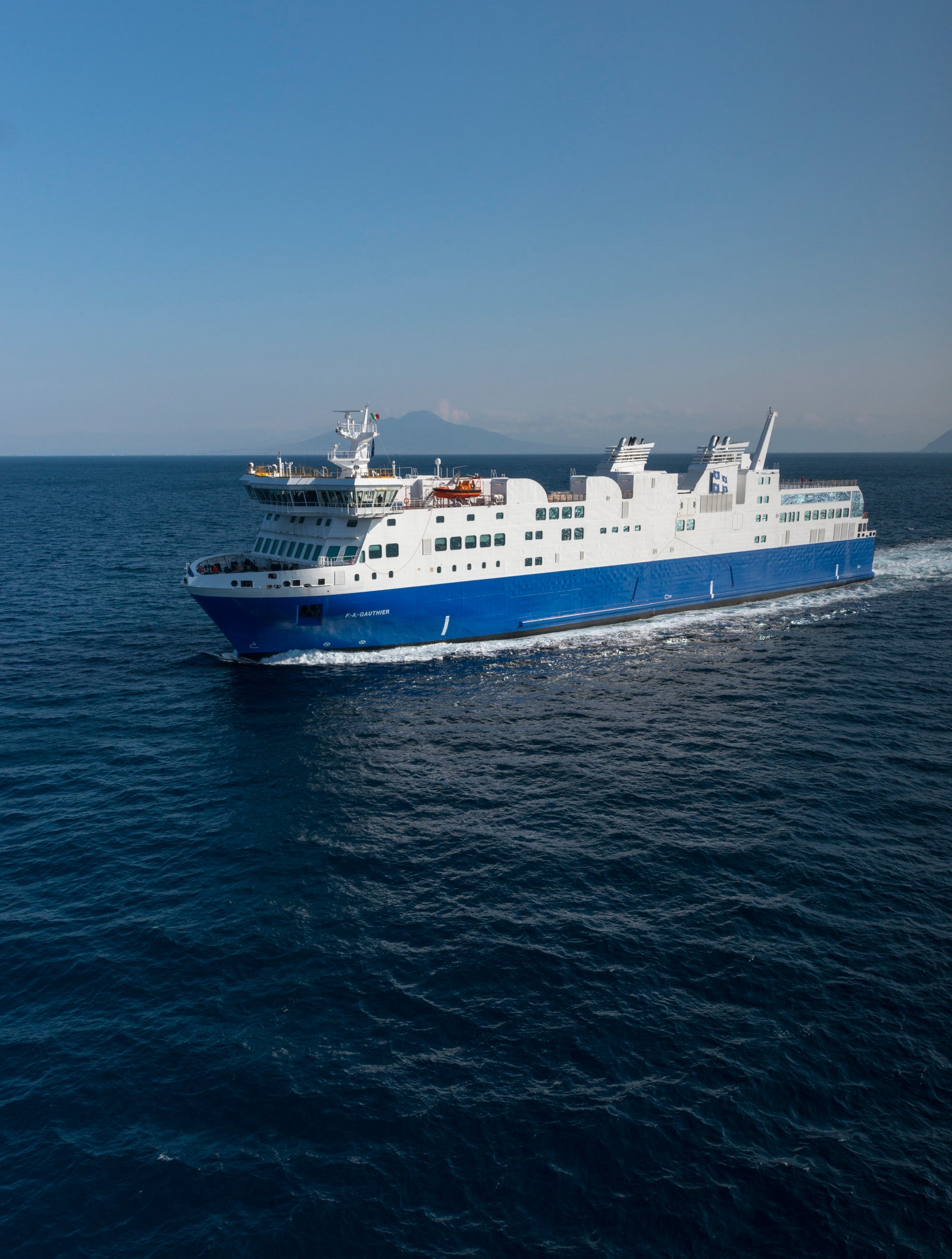 Fincantieri | Group - Business - Products - Ferries - Cruise FA Gauthier: Spacious blue and white "FA Gauthier" ferry in operation, multiple decks, visible lifeboats, cruising through open ocean, distant horizon and faint mountain silhouette in background, clear sky.