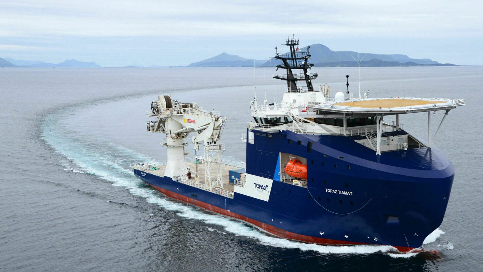 Fincantieri | Group - Business - Products - Offshore and Specialized Vessels - Energy - OFFSHORE SUBSEA CONSTRUCTIOn VESSEL - Topaz Tiamat: Large blue "Topaz Tiamat" offshore vessel in operation, helipad and visible maritime equipment, navigating calm open waters, "TOPAZ TIAMAT" text displayed on side, distant mountains, cloudy sky.