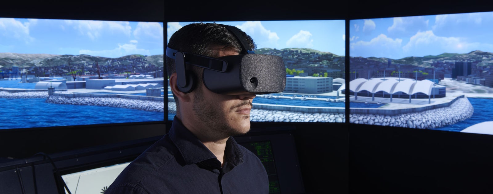 Fincantieri | People - Innovative Spirit: Man wearing a virtual reality headset immersed in a marine simulation, panoramic screen showing a coastal area, docks and buildings, numerical value 290.20 visible, technological or training environment.