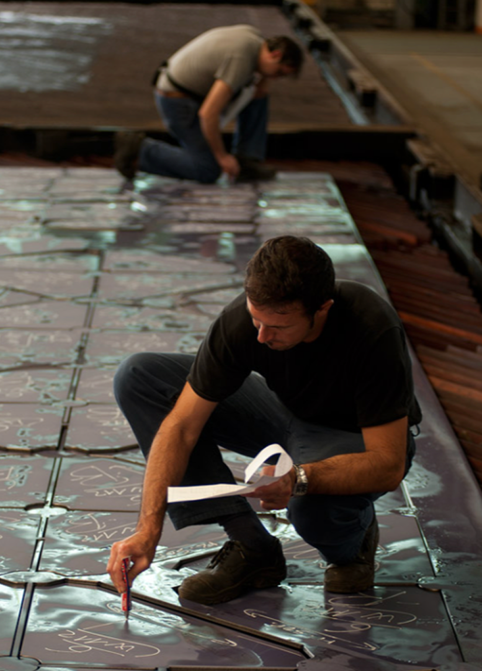 Fincantieri | Persone - Benessere Organizzativo: Due operatori osservano grandi lamiere metalliche in ambiente industriale, uno accovacciato con pennarello e blocco note, l’altro in secondo piano, superfici incise e riflettenti, contesto di fabbricazione.
