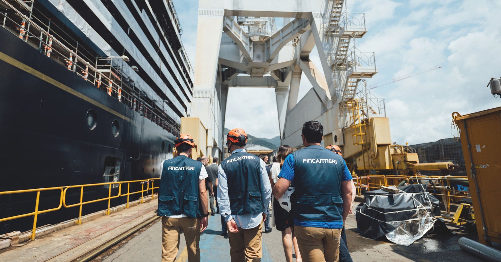 Fincantieri | Gruppo - Sostenibilità - Eccellenza industriale - Catena di fornitura: Gruppo di persone con giubbotti di sicurezza, testo ‘Fincantieri’ in evidenza, che passeggiano in cantiere navale all’aperto con attrezzatura industriale e nave ormeggiata, cielo sereno e montagne sullo sfondo.